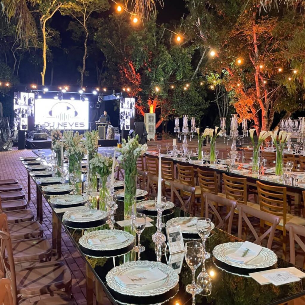 Decoração de jantar de luxo ao ar livre no Restaurante Mata Nativa, com mesas longas de vidro, flores brancas, iluminação de varal de luzes e palco com DJ ao fundo em meio às árvores.