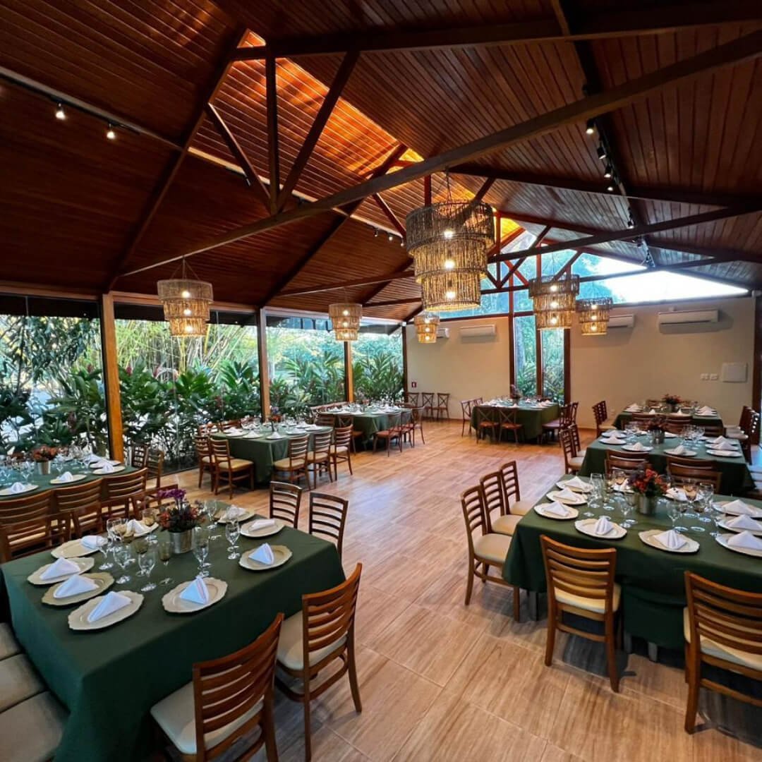 Salão interno climatizado do Restaurante Mata Nativa com paredes de vidro, vista para o jardim, mesas postas com toalhas verdes, cadeiras de madeira e lustres artesanais elegantes.