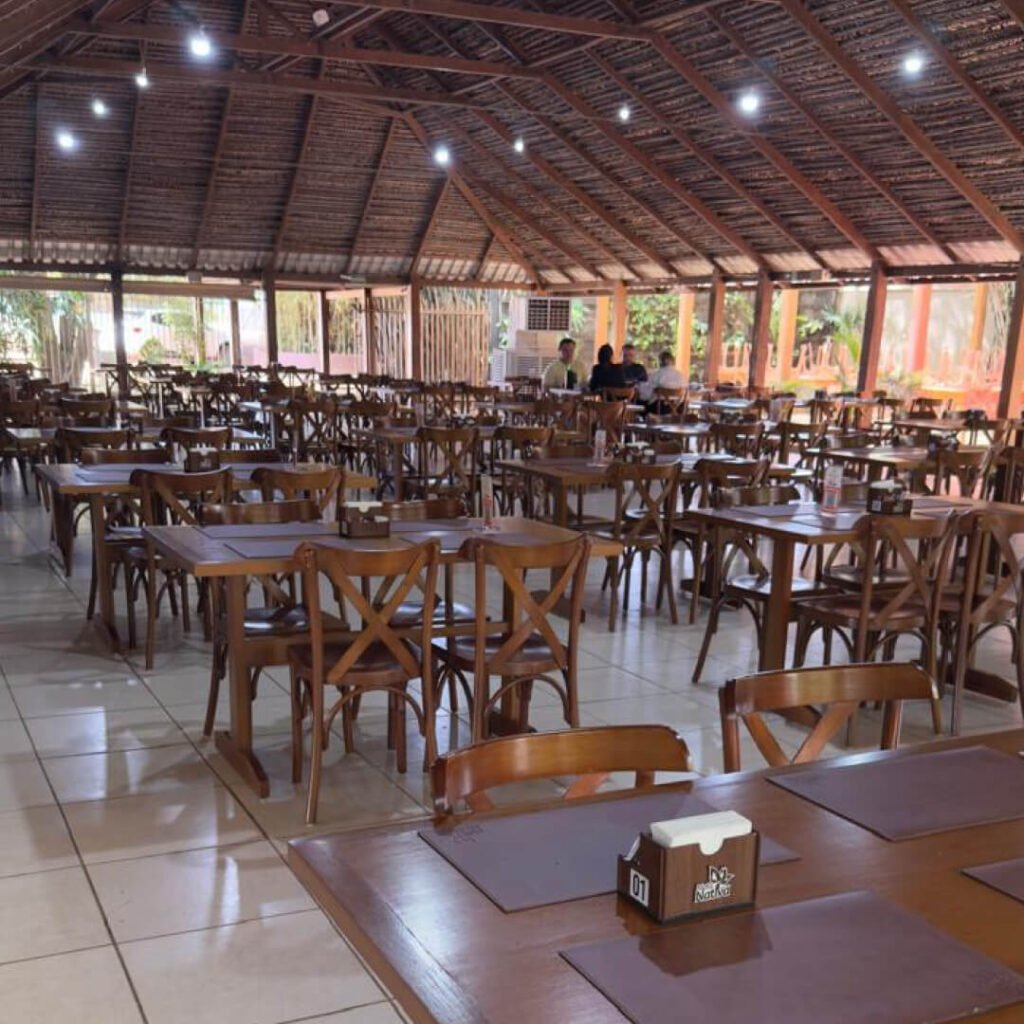 Vista interna do amplo salão do Restaurante Mata Nativa, com mesas e cadeiras de madeira organizada sob um teto alto de estrutura rústica e palha, em ambiente arejado.